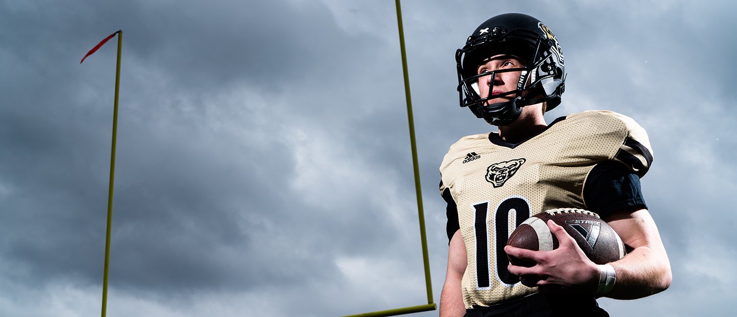 A man in an Oakland University jersey and helmet, holding a football.