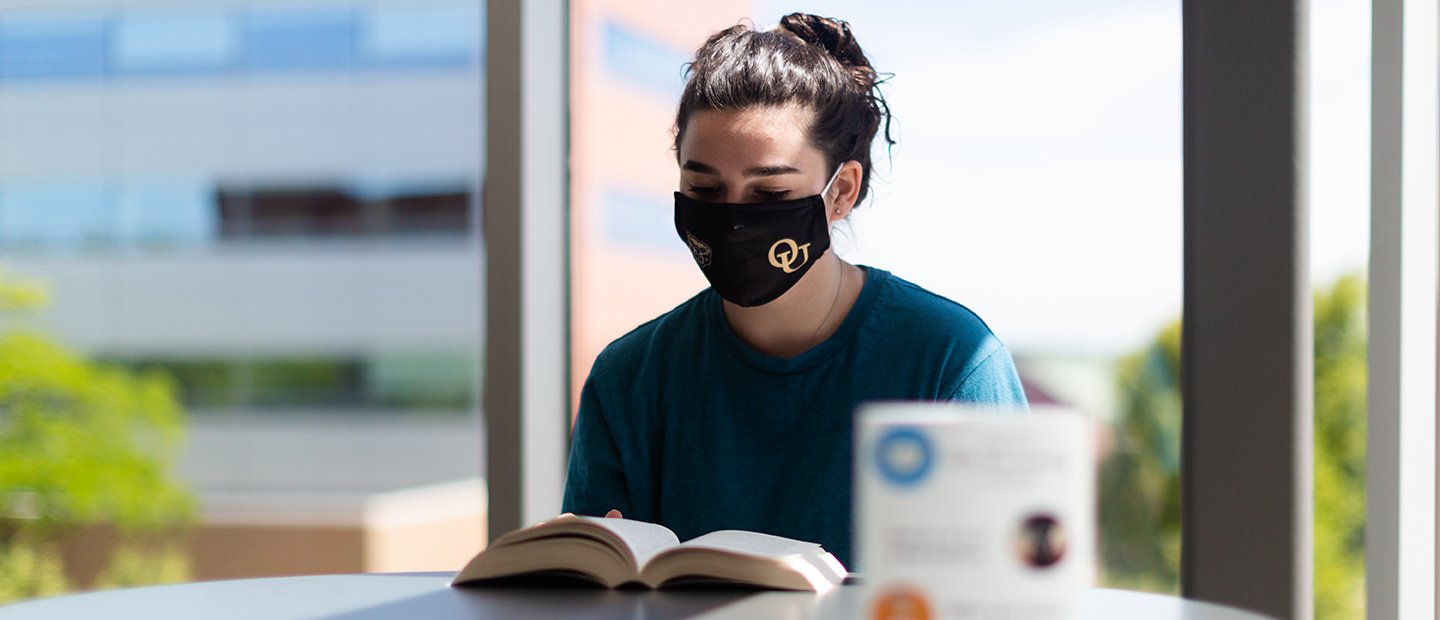 An Oakland university student wearing a face mask, reading a book at a table.