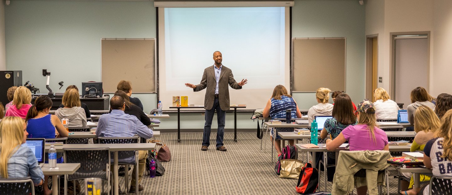 A professor standing at the front of a full classroom, talking and gesturing with his hands.