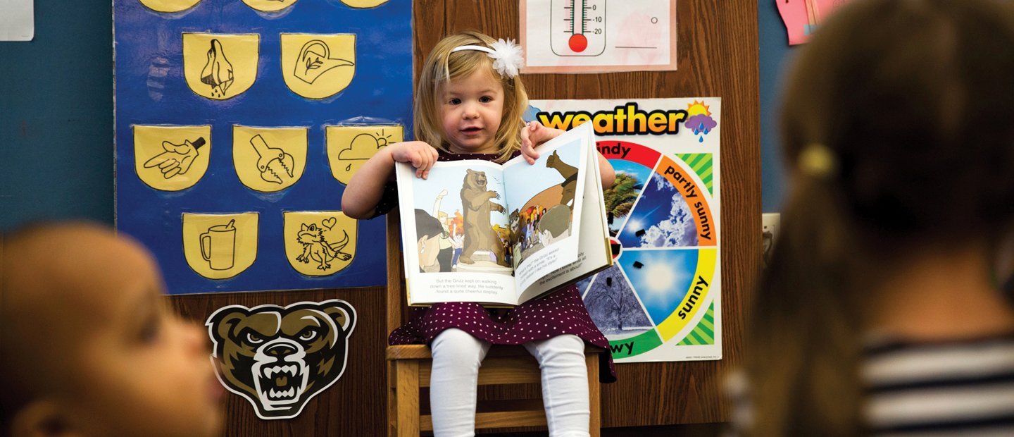 A young girl seated at the front of a classroom, showing a book to the other students.