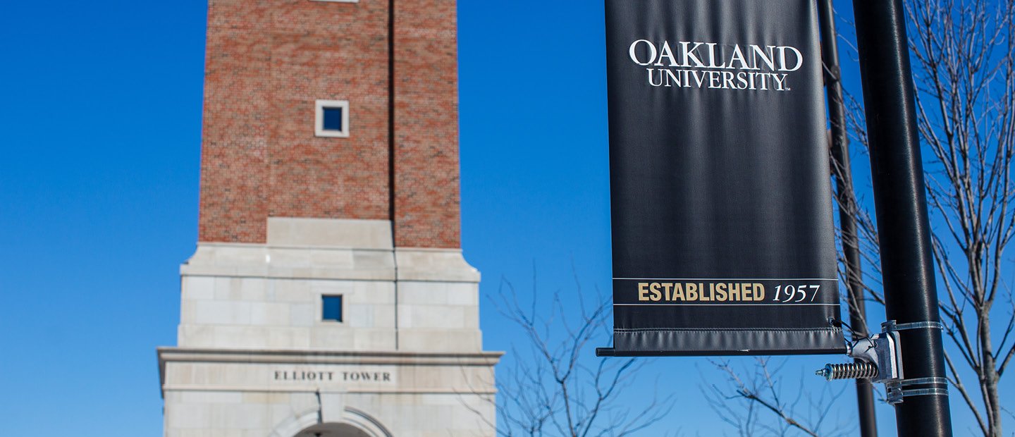 A black Oakland University banner on a post, in front of Elliott Tower.