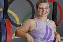 image of Kate Nye standing in a weight room with the Olympics symbol behind her