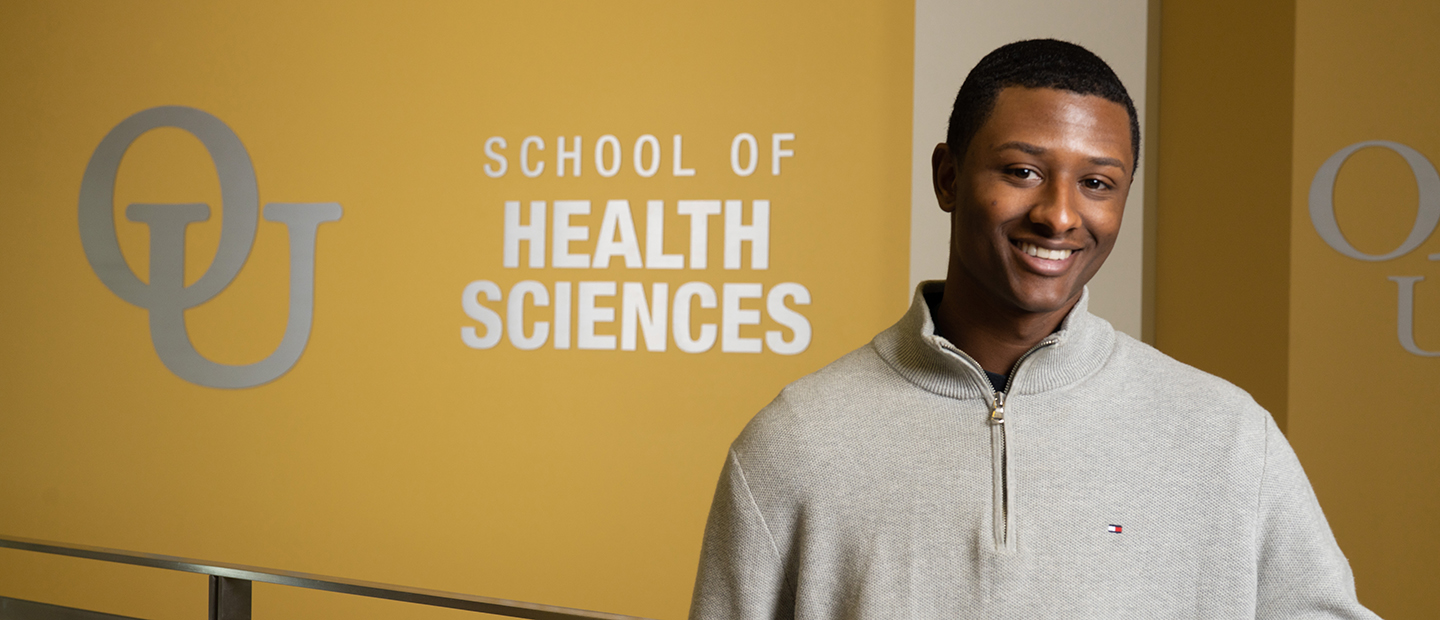 image of a student standing in the Human Health Building in front of a sign with the interlocking OU and School of Health Sciences text