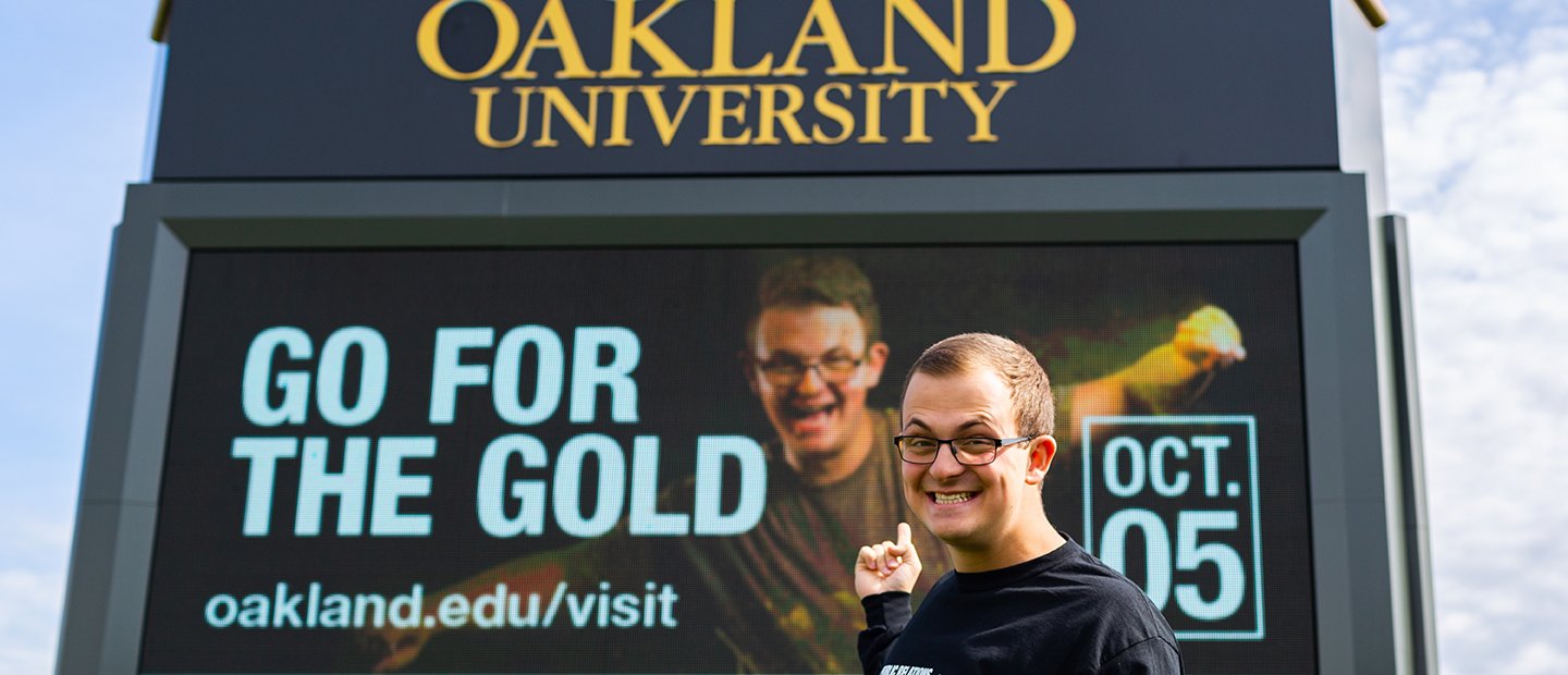 Young man pointing to an ad that he is featured in on an Oakland University electronic marquee.