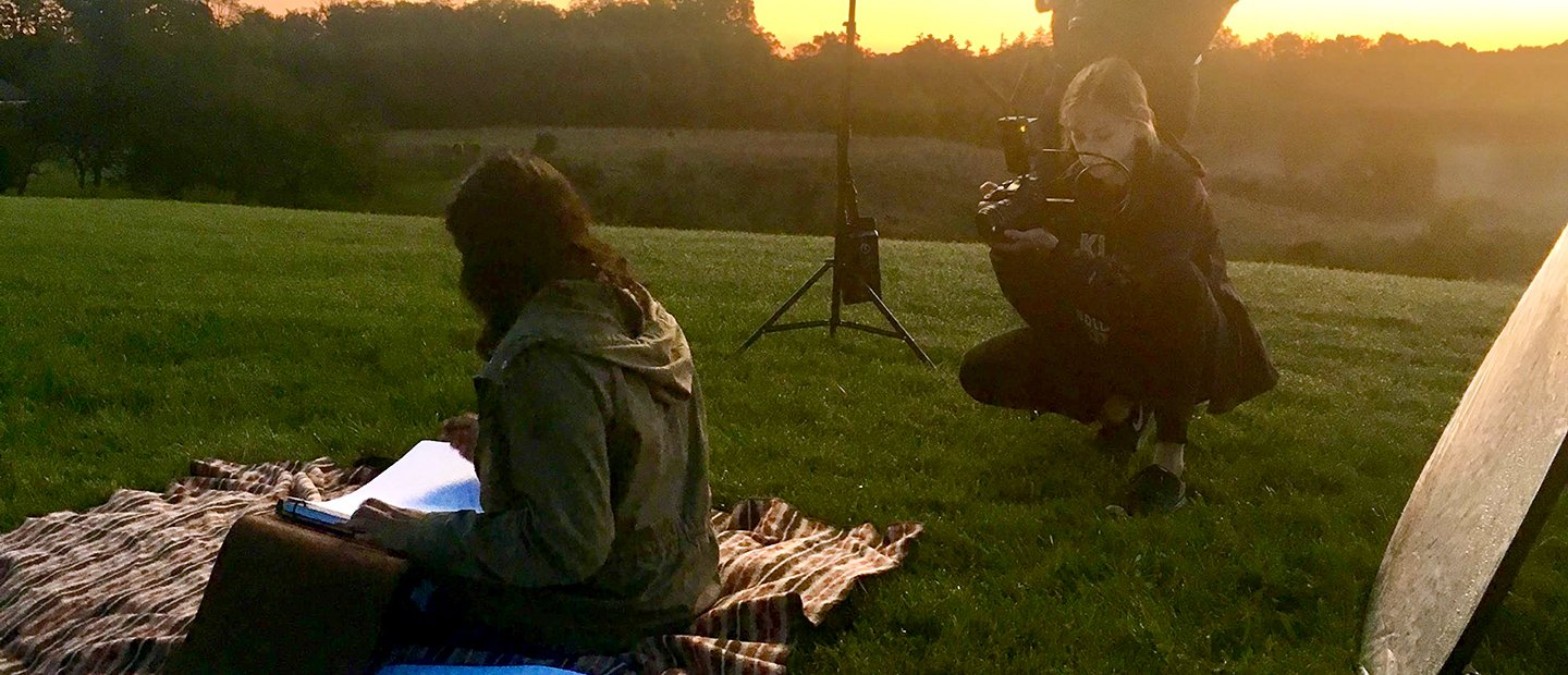 A young woman taking photographs of a person seated on a blanket outside, reading a book.