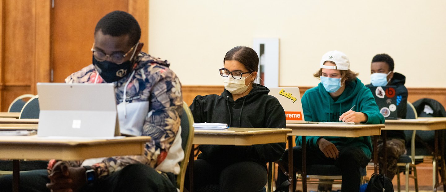 Undergraduate Catalog Four students sitting at individual desks in a row, all wearing face masks.