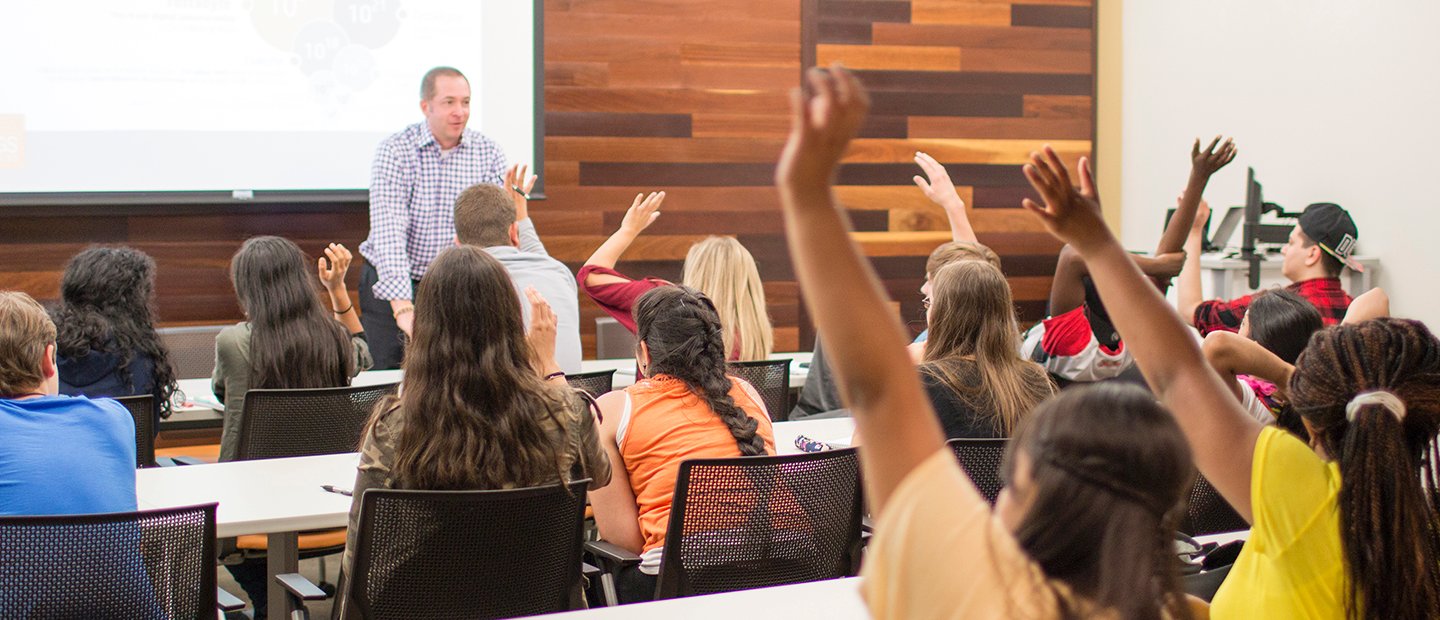 A room full of students in a classroom, many of them raising their hands.