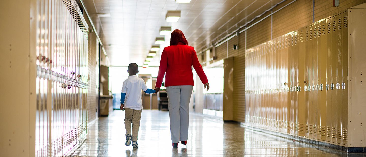 An adult and a child holding hands, walking down a hallway full of lockers. 