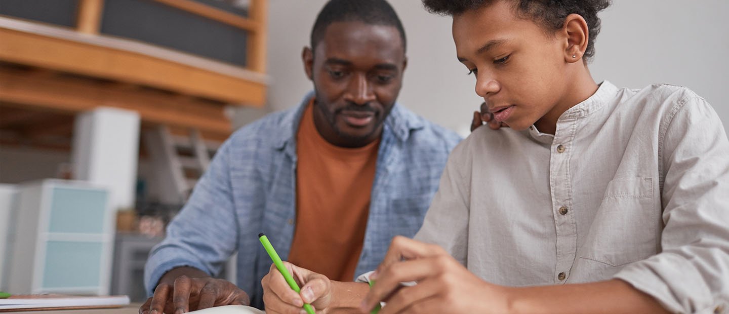 A parent looking over his son's shoulder as he writes in a notebook.