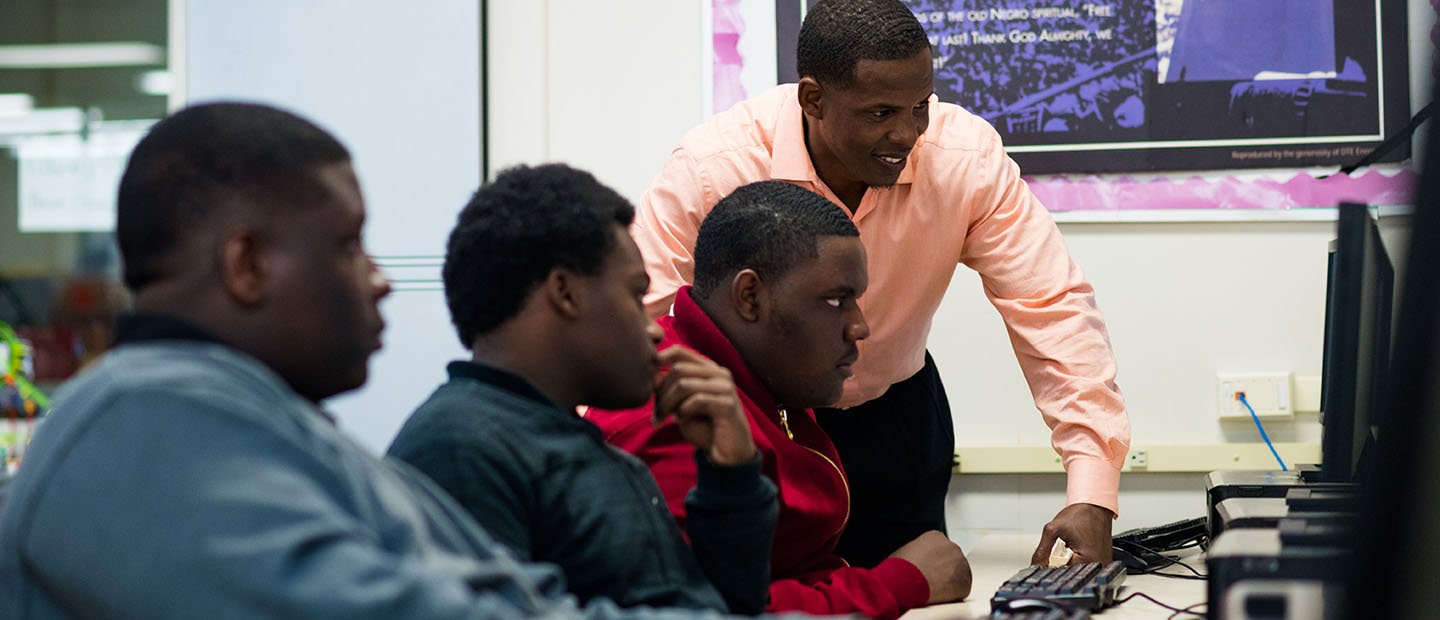 A man standing over a row of three young men working at computers.