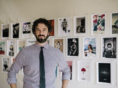 Man with hands on hips standing in front of three rows of artwork hanging on a brick wall