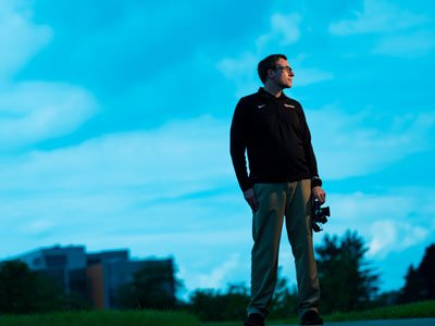 Man standing outside on a sidewalk at dusk with a camera in his hand.