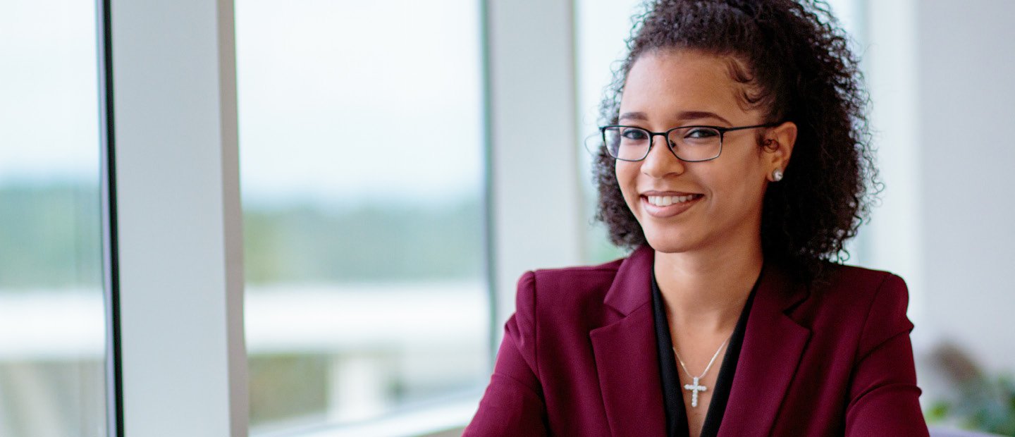 A woman in a dark red jacket, seated in front of a window, smiling at the camera.