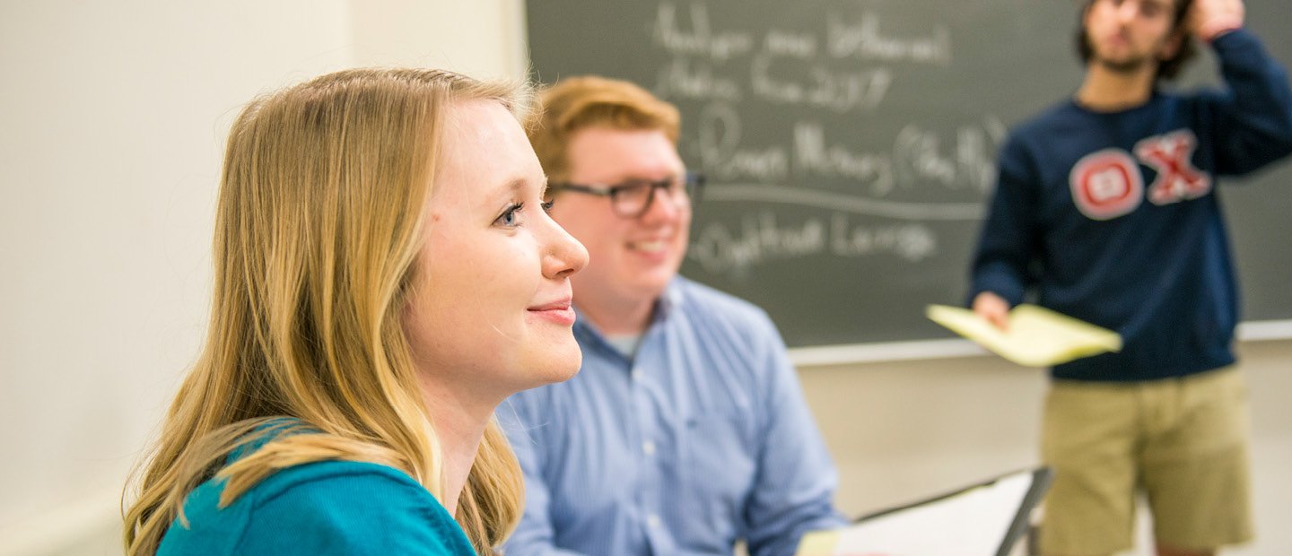 A young man and woman seated, looking towards the front a classroom while a young man stands behind them by a chalkboard.