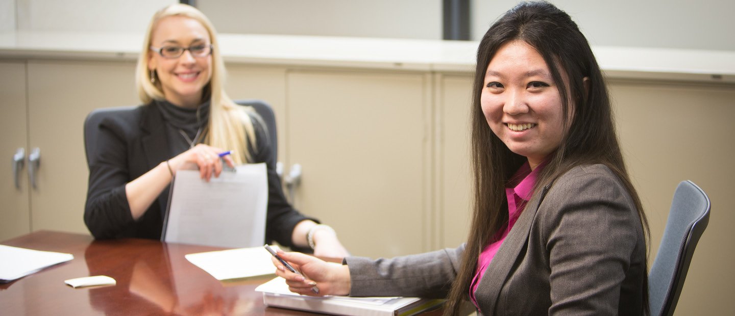 Two women seated a a table with notebooks and pens, smiling at the camera.