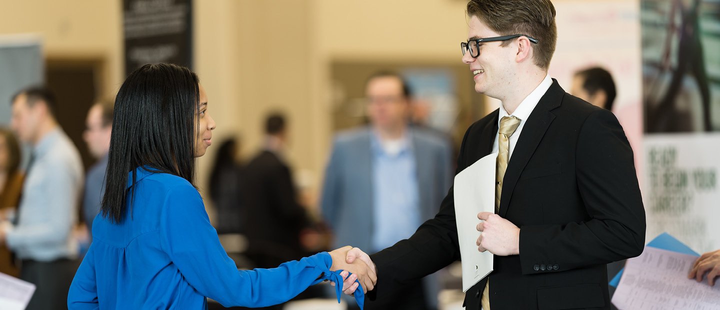 A young man in a suit holding a document, shaking a young woman's hand.