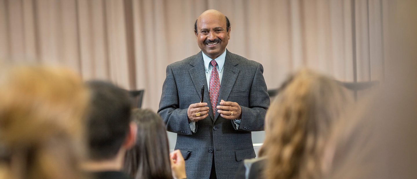 A professor in a suit, facing the camera, smiling at a room full of students.