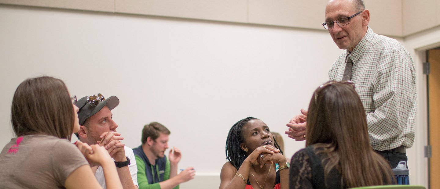 A professor standing next to a table with four students, talking to them.