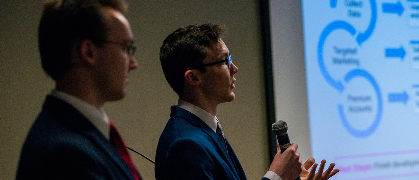 Two young men in suits, one holding a microphone, standing next to a projector screen.