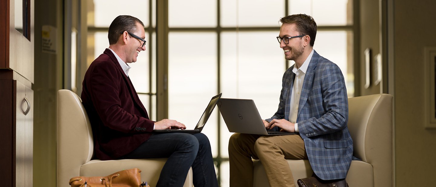 Two men seated in leather chairs with laptops, facing each other.