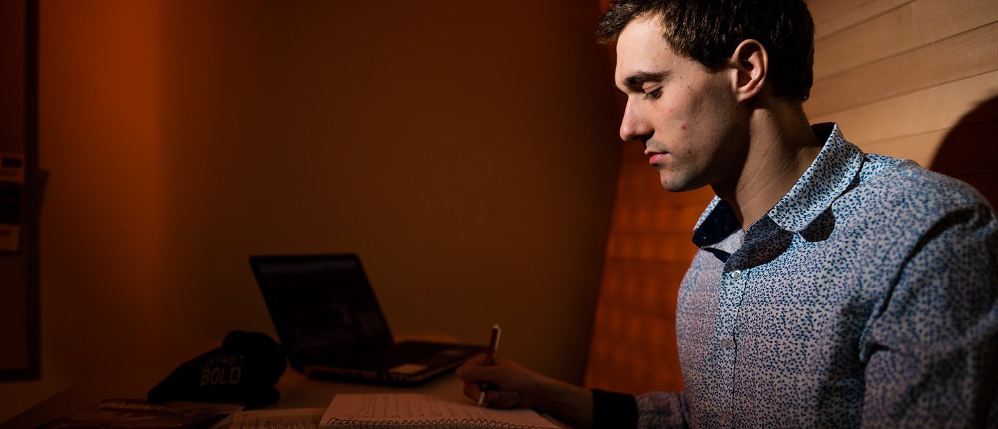 A man sitting in a booth, writing in a notebook with an open laptop next to him.