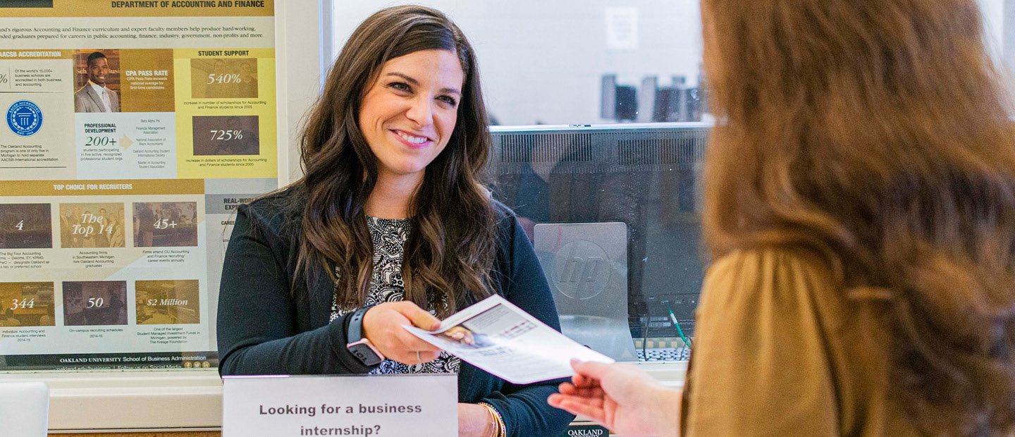 A woman handing a flyer to another person A woman standing in front of a graphic poster of statistics, handing a flyer to another person.