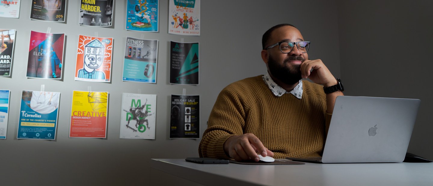 A man seated at a desk with a laptop and a wall of colorful posters behind him.