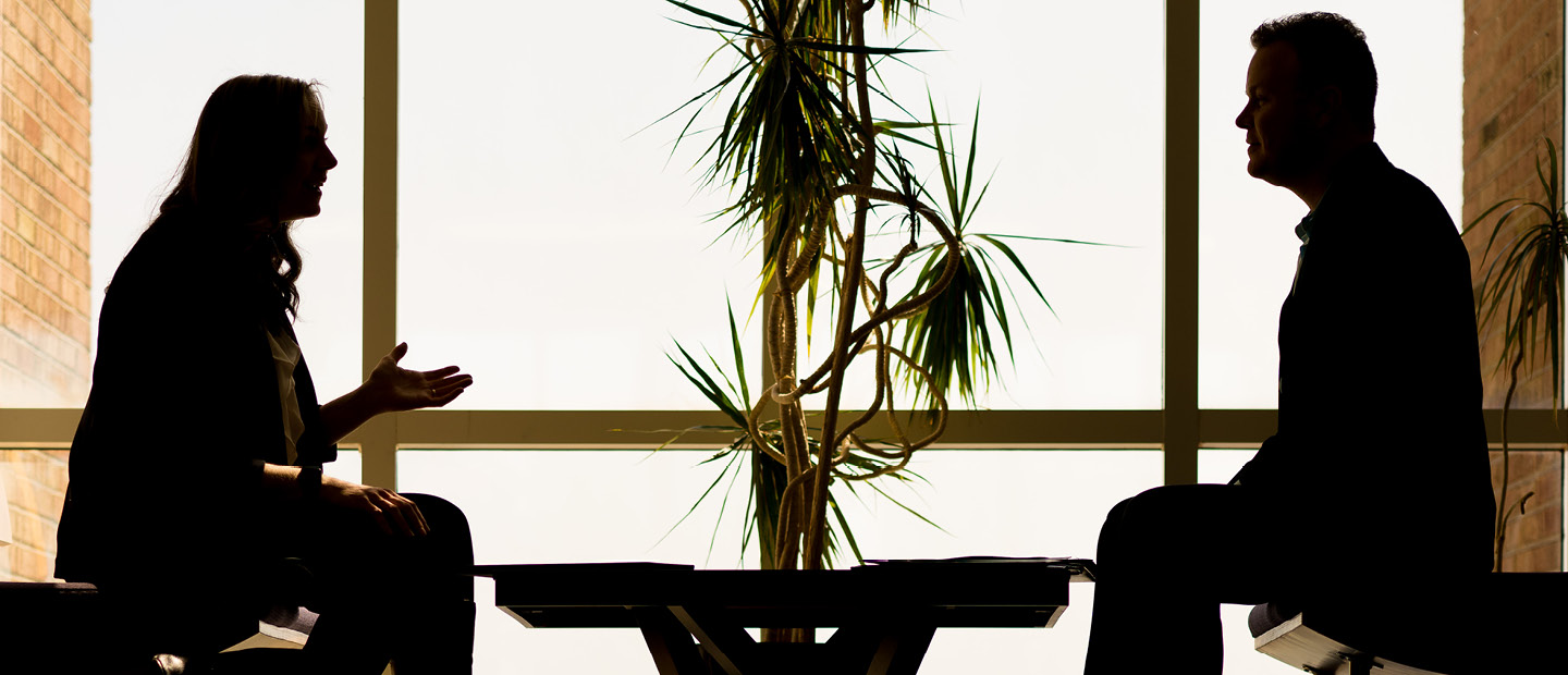 The silhouette of two people seated across a table from each other, talking.