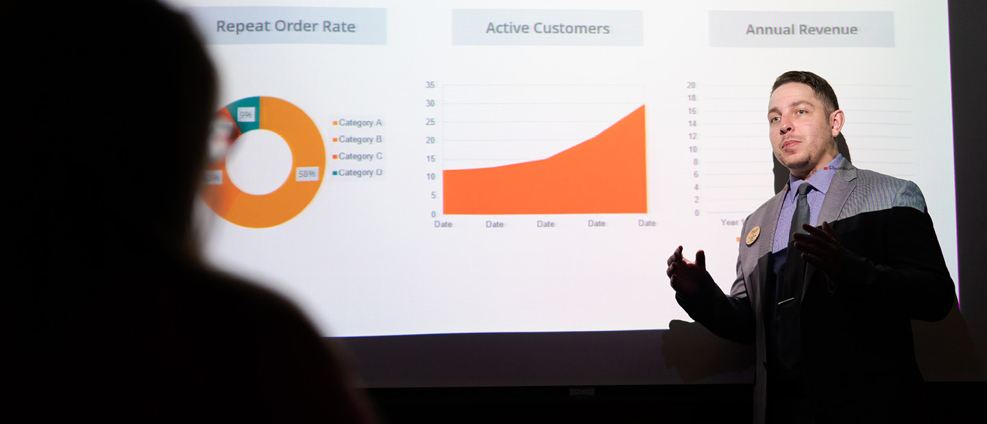 A man in a suit presenting in front of a projector screen showing colored charts and graphs.