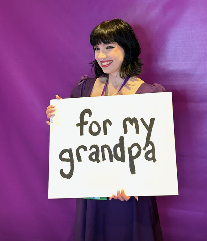 Sophia Vecchi holds up a sign that reads for my grandpa in front of a purple background.