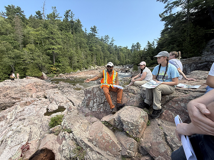 Students listen to an expert discuss different rocks as they sit on rocks at Chippewa Falls.