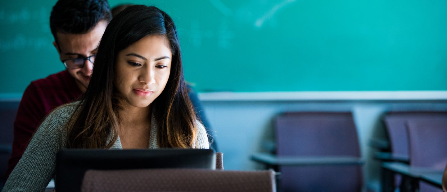 two students seated, looking at laptops, with a green chalkboard in the background