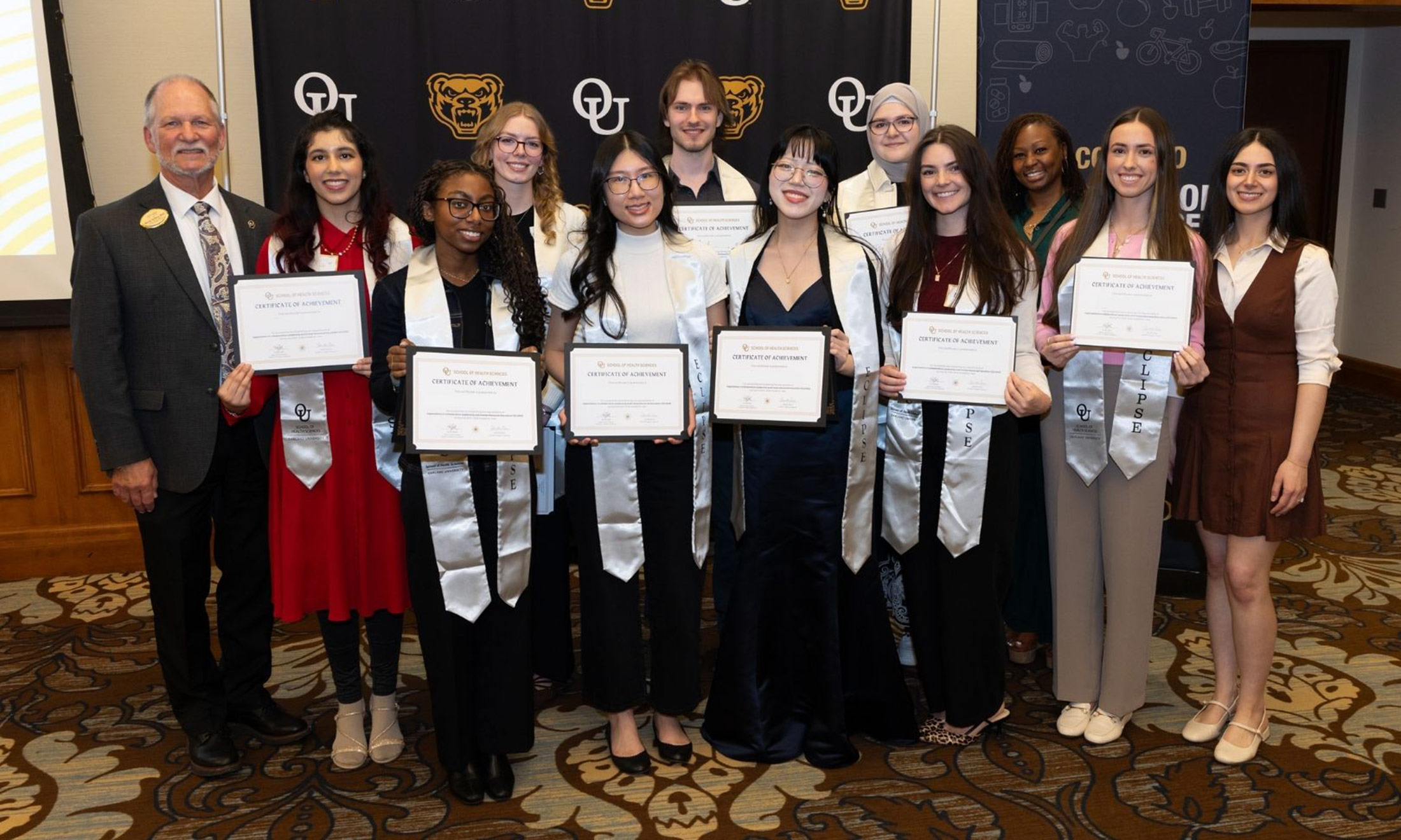A group of students hold certificates in front of an Oakland University banner