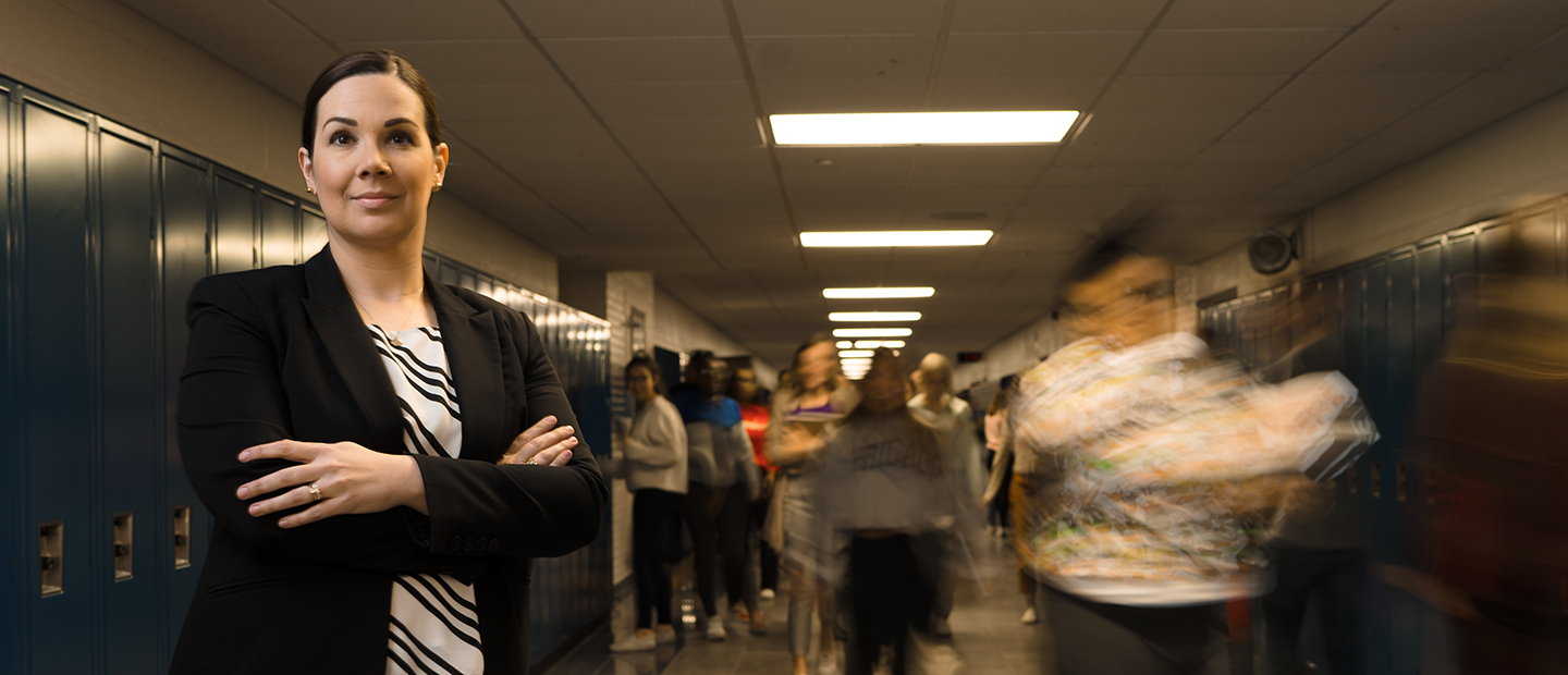 A woman standing with her arms folded across her chest, in a hallway of a school with blue lockers along the wall and people walking behind her.