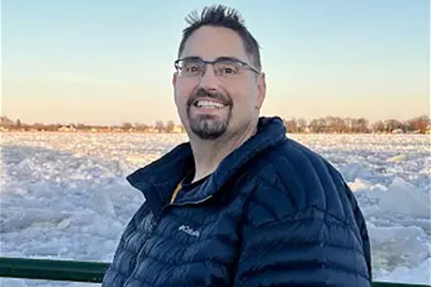A man facing the camera. He is outside and there is snow behind him.