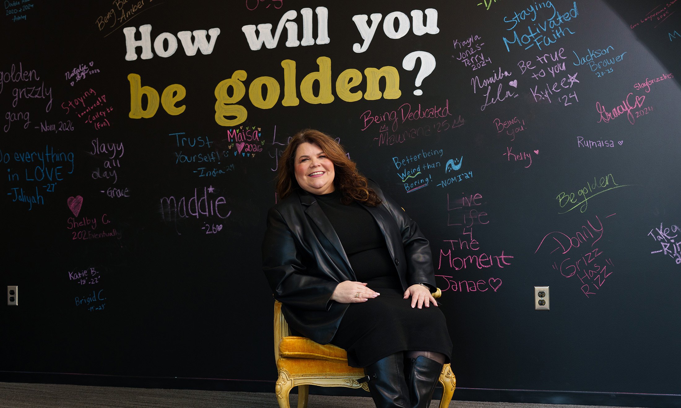 A woman sitting in a gold seat in front of a chalkboard that reads 