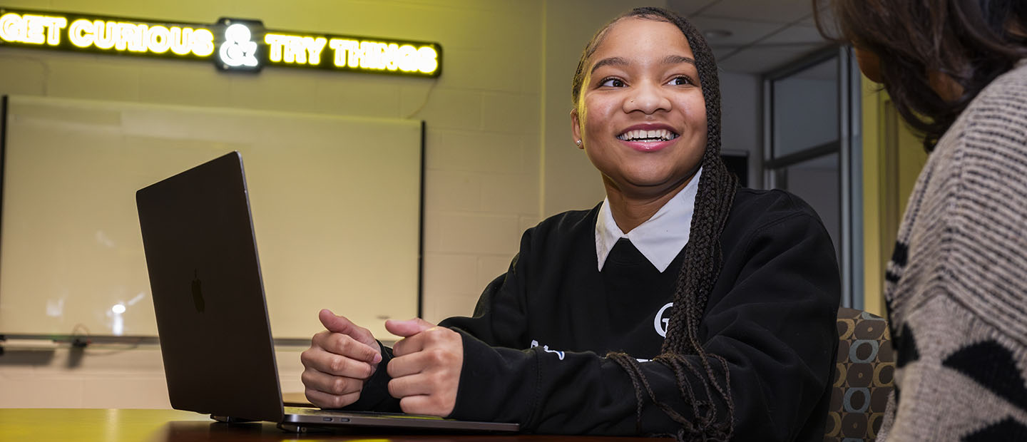 A student seated with a laptop, talking to another person