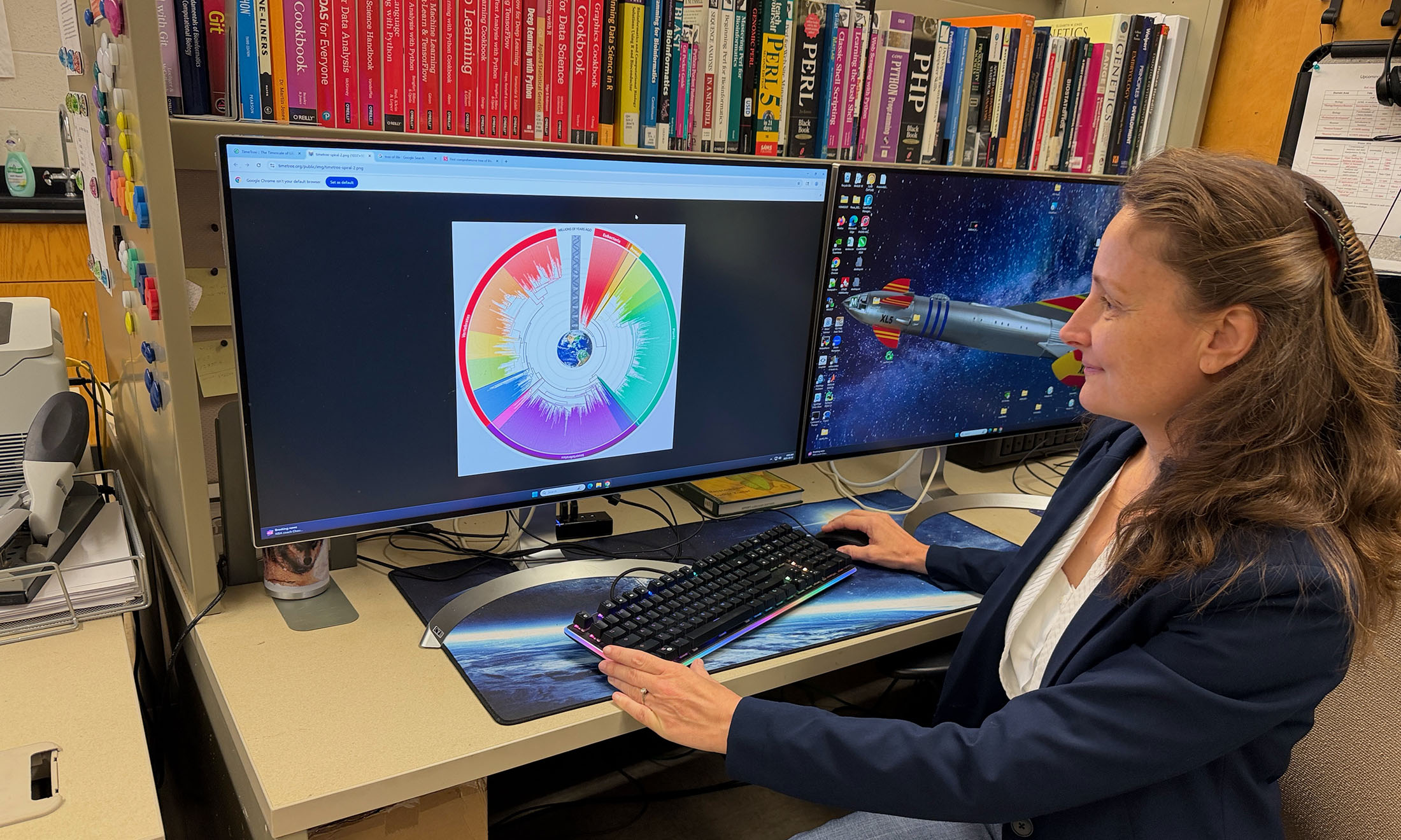 a professor sitting by a desktop computer; genomic trees display on the screen