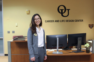 woman smiling at camera in front of a desk with OU on the wall behind her