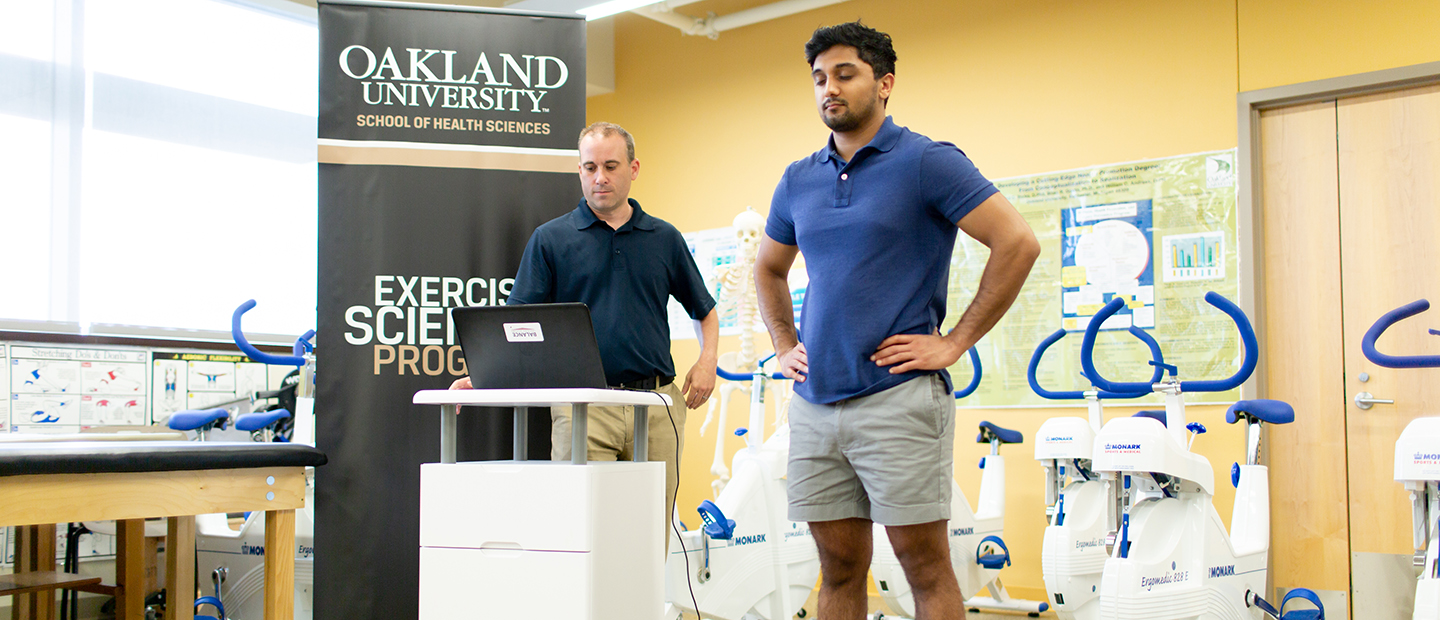 A man standing on a scale while another man reads data on a computer screen, in a fitness studio.