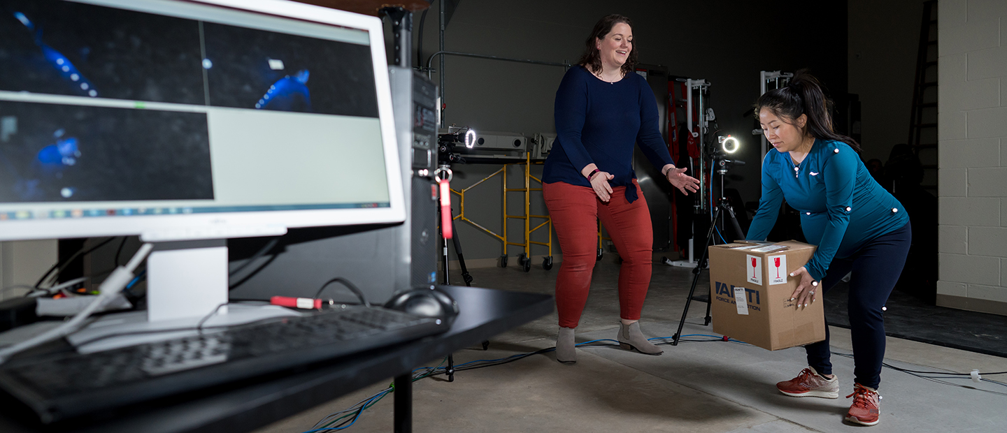 A woman with sensors attached to her body lifts a box, as another woman watches her and demonstrates the motion.