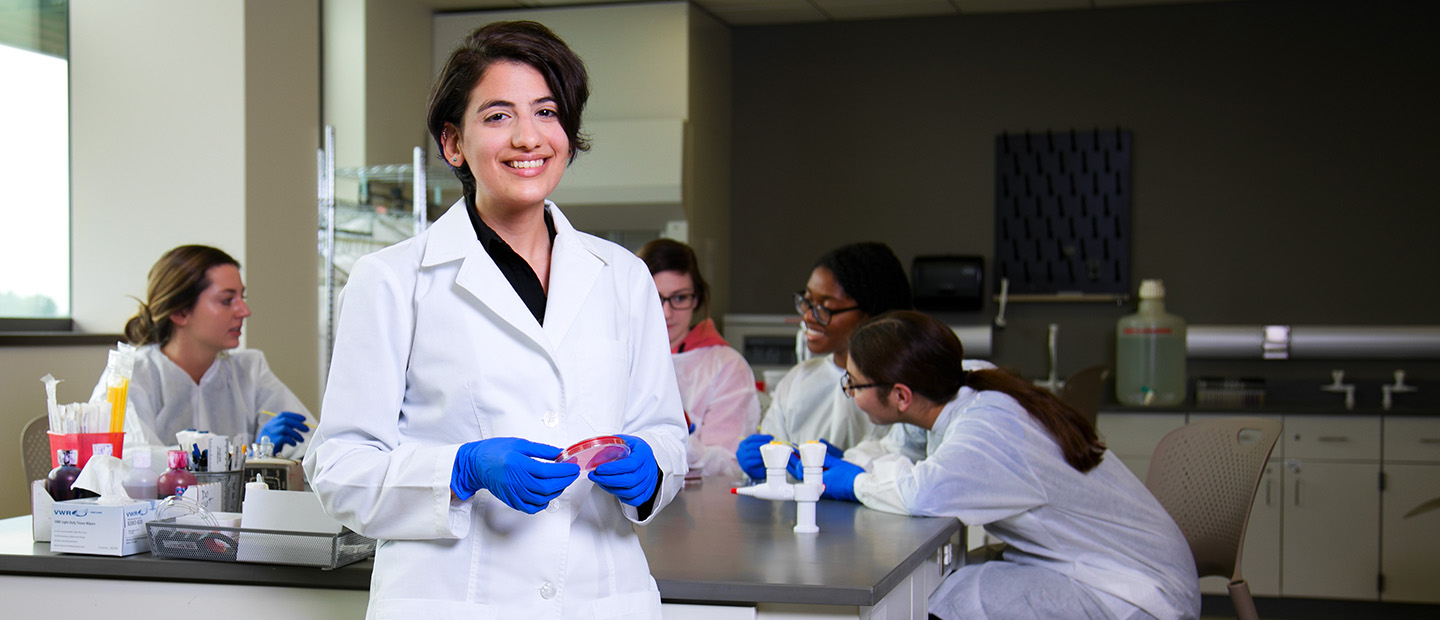 young woman in a white lab coat and blue gloves, smiling at the camera