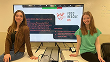 Oakland University students, Sarah Nyquist and Rachel Whalen, seated in front of a large monitor displaying information about Food Rescue US.