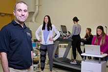 A man smiling at the camera while a woman walks on a treadmill and others perform fitness evaluations.
