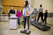 A woman walking on a treadmill, another woman standing on a scale, while students perform fitness evaluations.