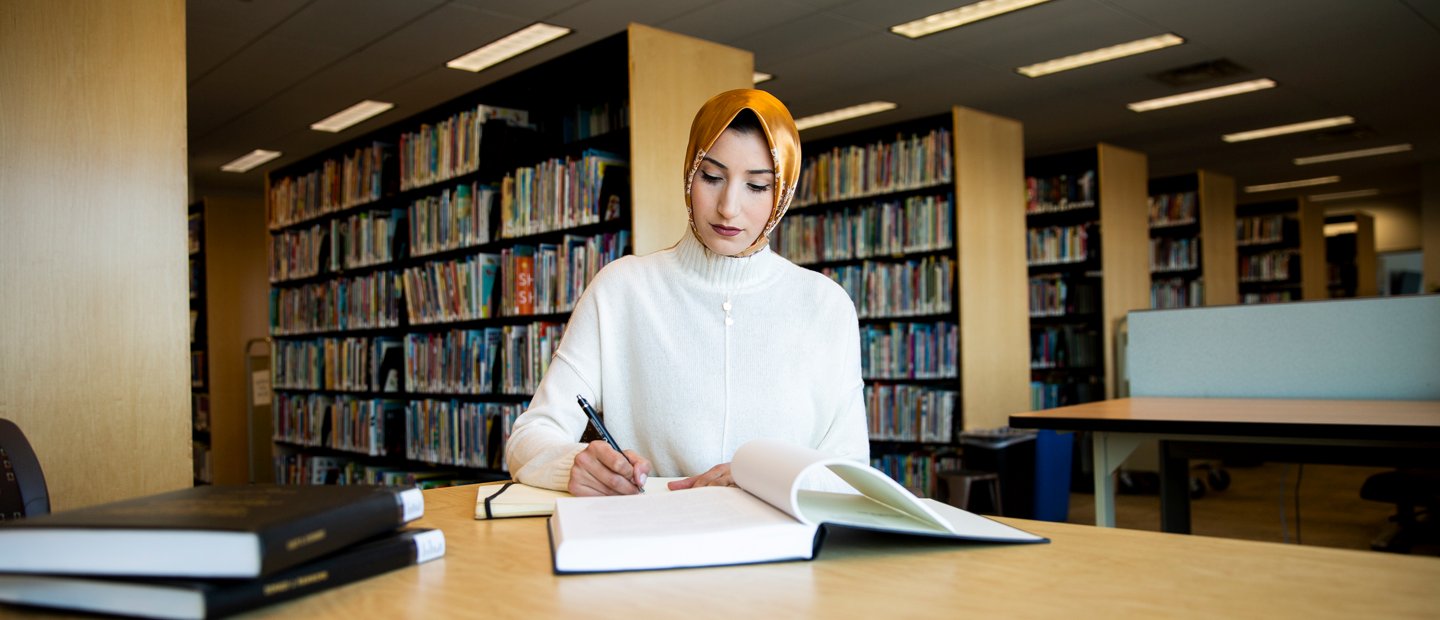 A woman seated at a table with open books, with rows of bookshelves behind her.