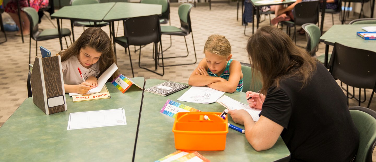 Young students sitting with a woman at a table, writing and reading Two young girls sitting with a woman at a green table, writing and reading.