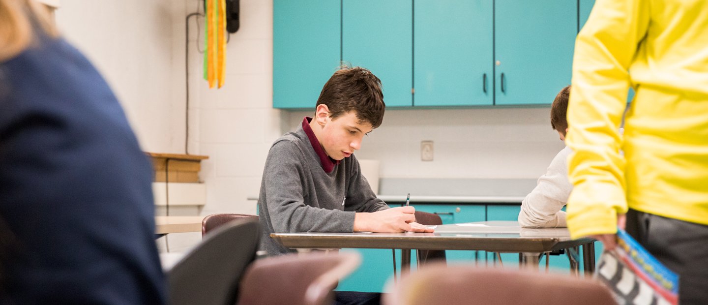 A boy seated at a table in a classroom, writing A boy seated at a table in a classroom, writing.