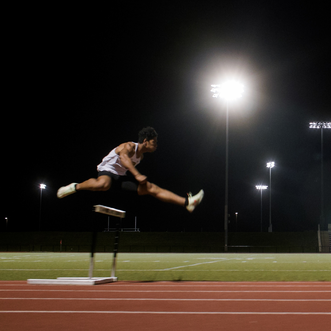 A track runner jumping over a hurdle at night
