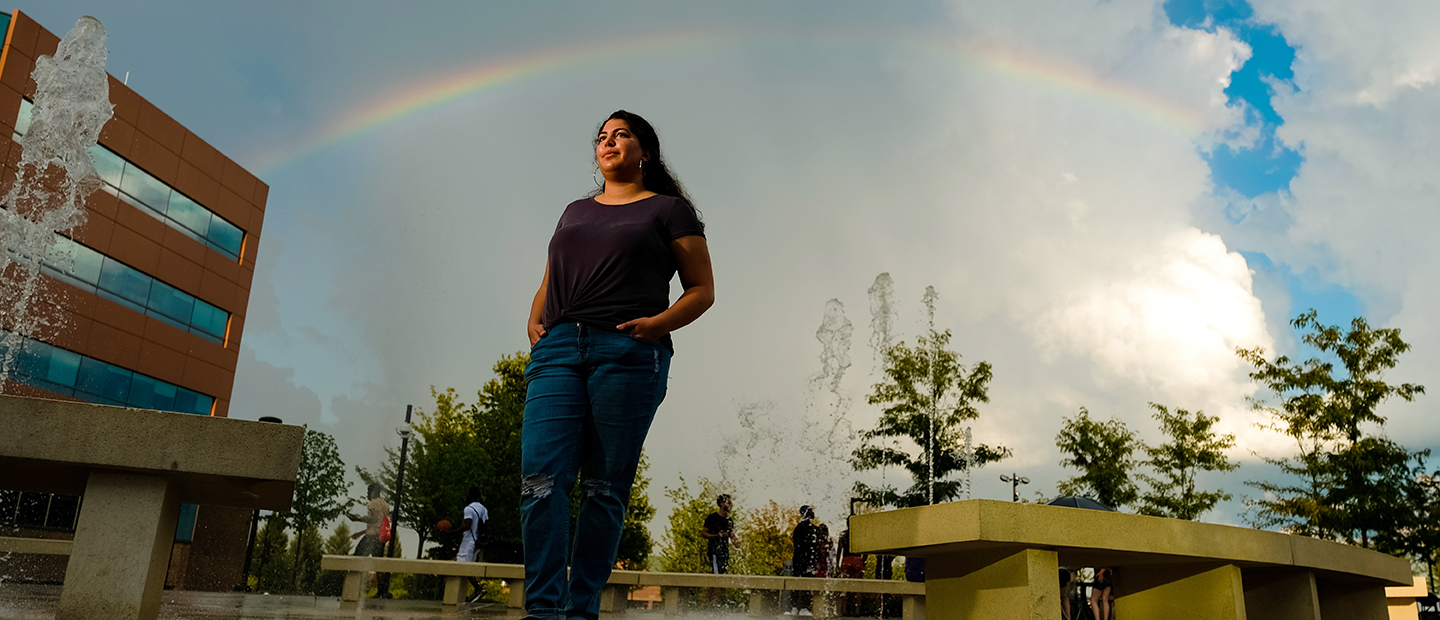 A young woman standing with her hands in her pockets on Oakland University's campus, with a rainbow and fountain behind her.
